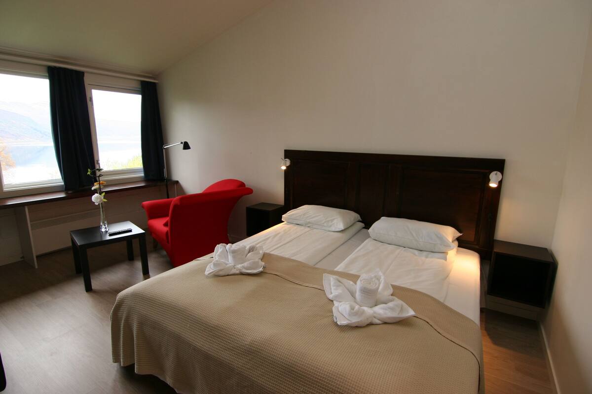 Quarto de hotel com cama, uma cadeira vermelha, mesa de cabeceira e janela com cortinas.