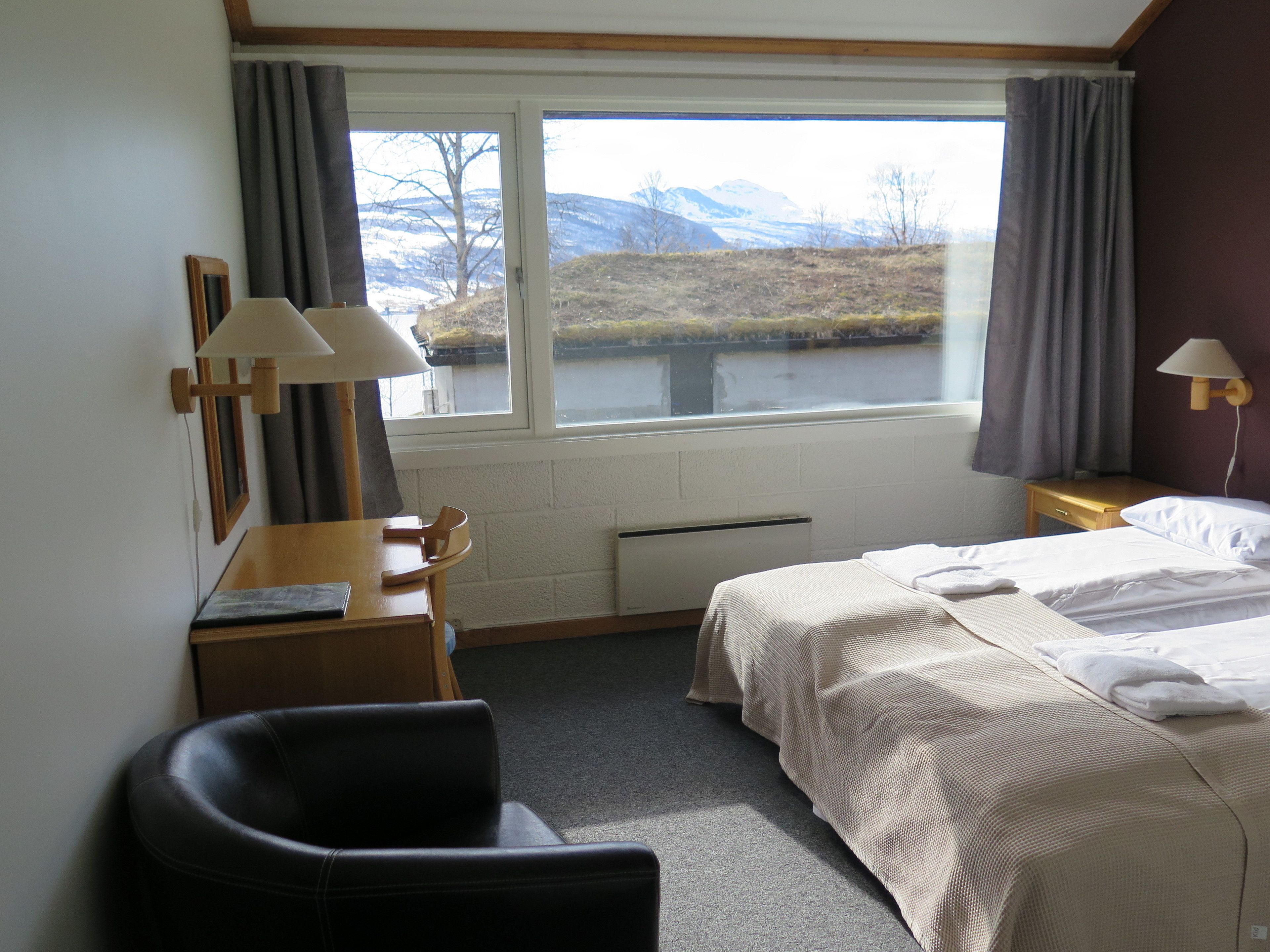 Une chambre d’hôtel avec deux lits, un bureau, une chaise et une fenêtre donnant sur des montagnes et un lac.