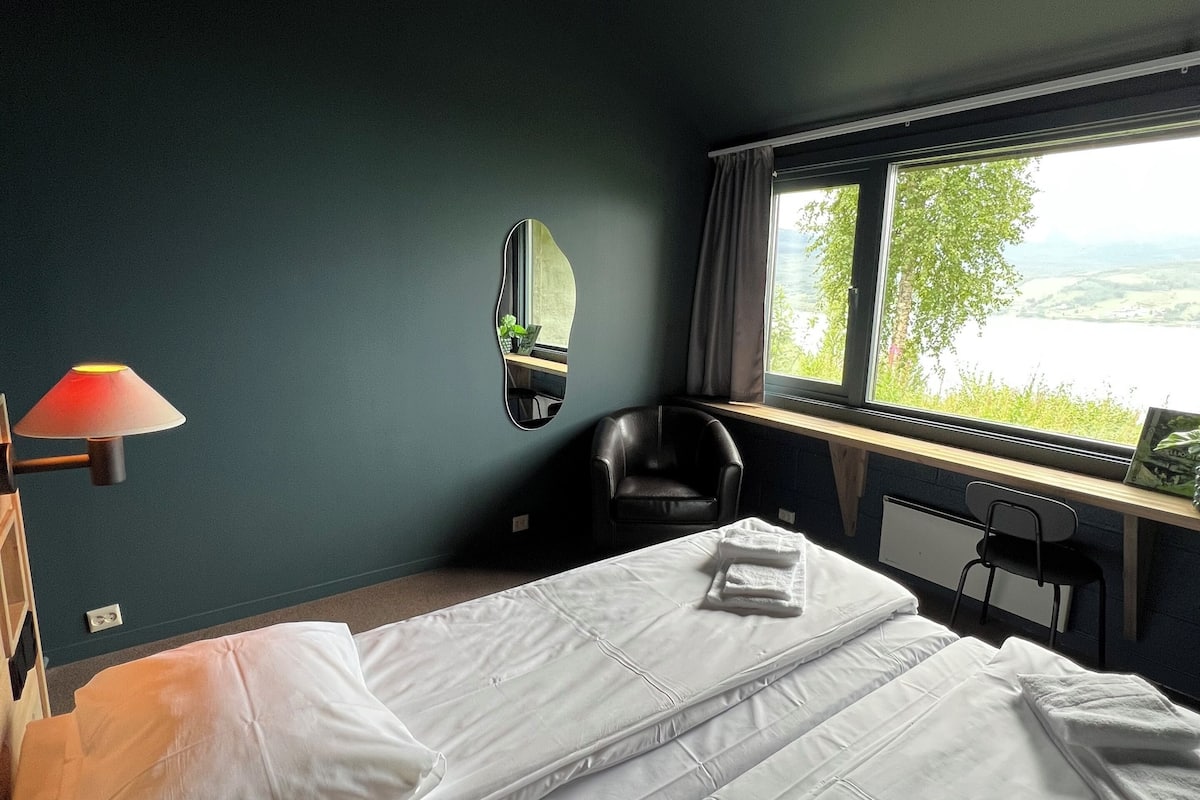 Quarto de hotel com cama, escrivaninha, cadeira, abajur, espelho e janela com vista para o verde.