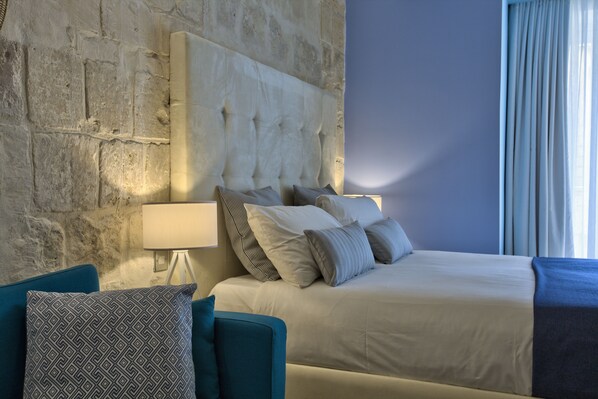 A hotel room with a stone wall, a large bed with multiple pillows, a bedside table with a lamp, and a blue armchair.