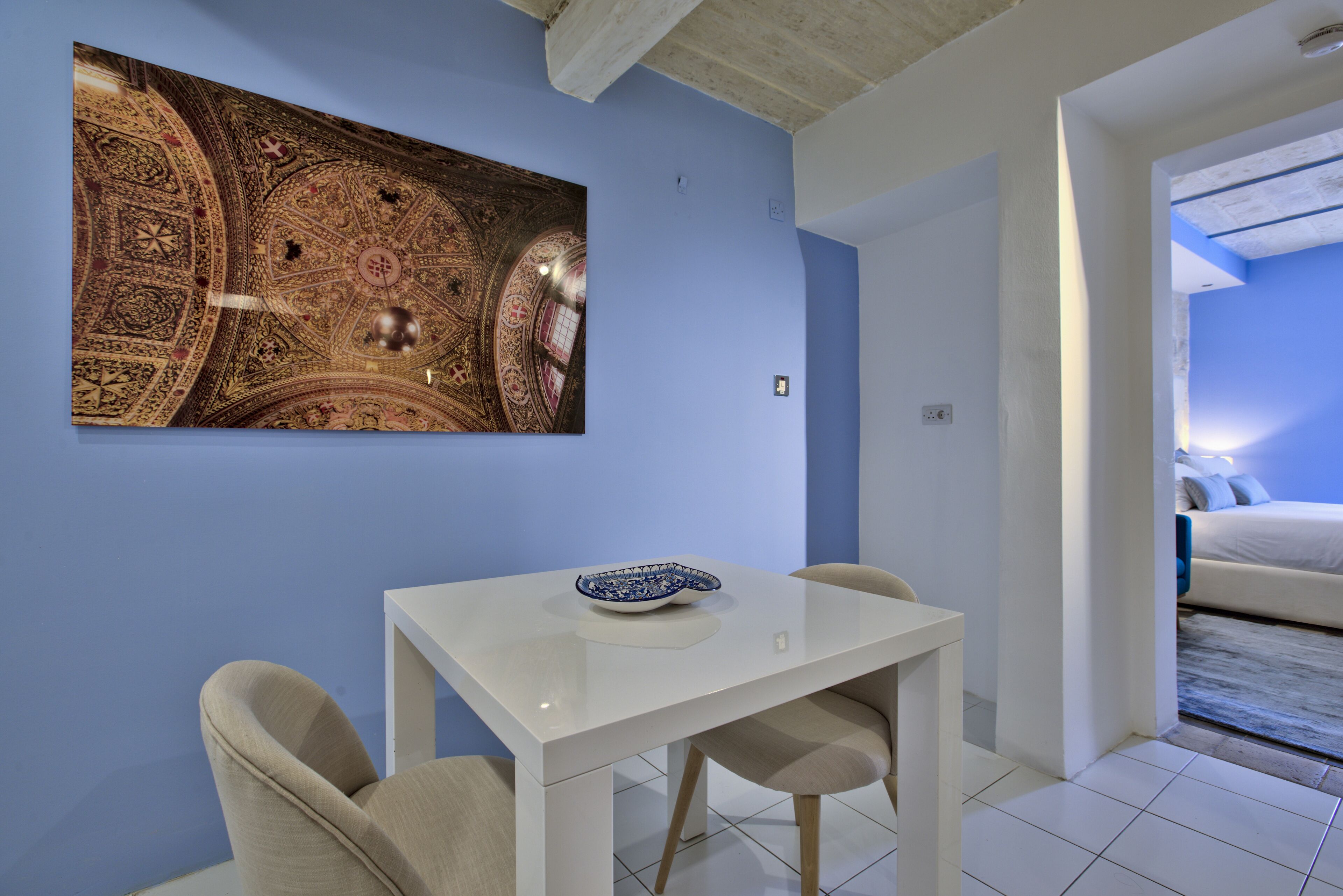 A modern room with a blue wall, a white table with two chairs, a large decorative ceiling, and a bed with blue bedding.