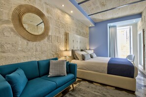 A bedroom with a stone wall, a round mirror, a blue sofa, a bed with multiple pillows, and a view through a window.