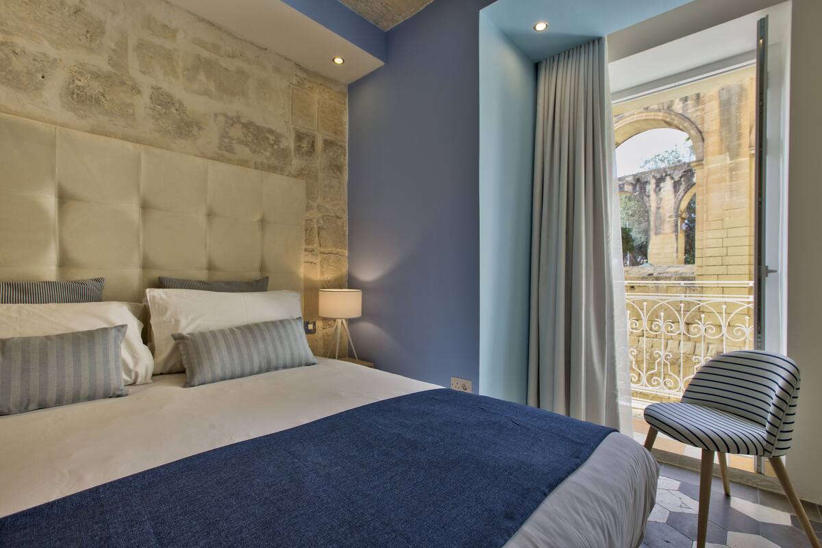 A hotel room with a large bed, a view of an outdoor balcony, and a stone wall.