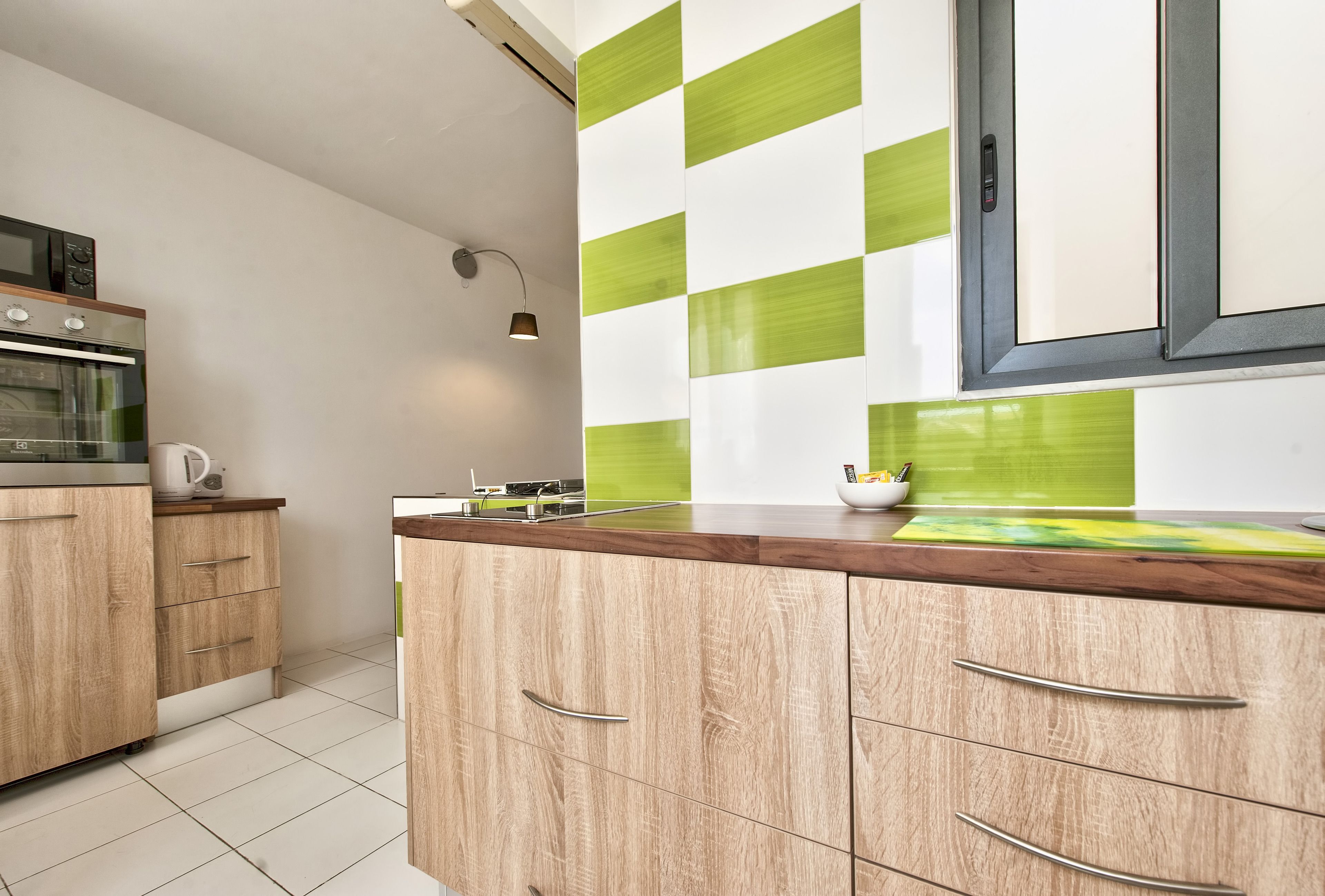 Een moderne keuken met houten kastjes, een magnetron, een oven en een backsplash met groene en witte tegels.