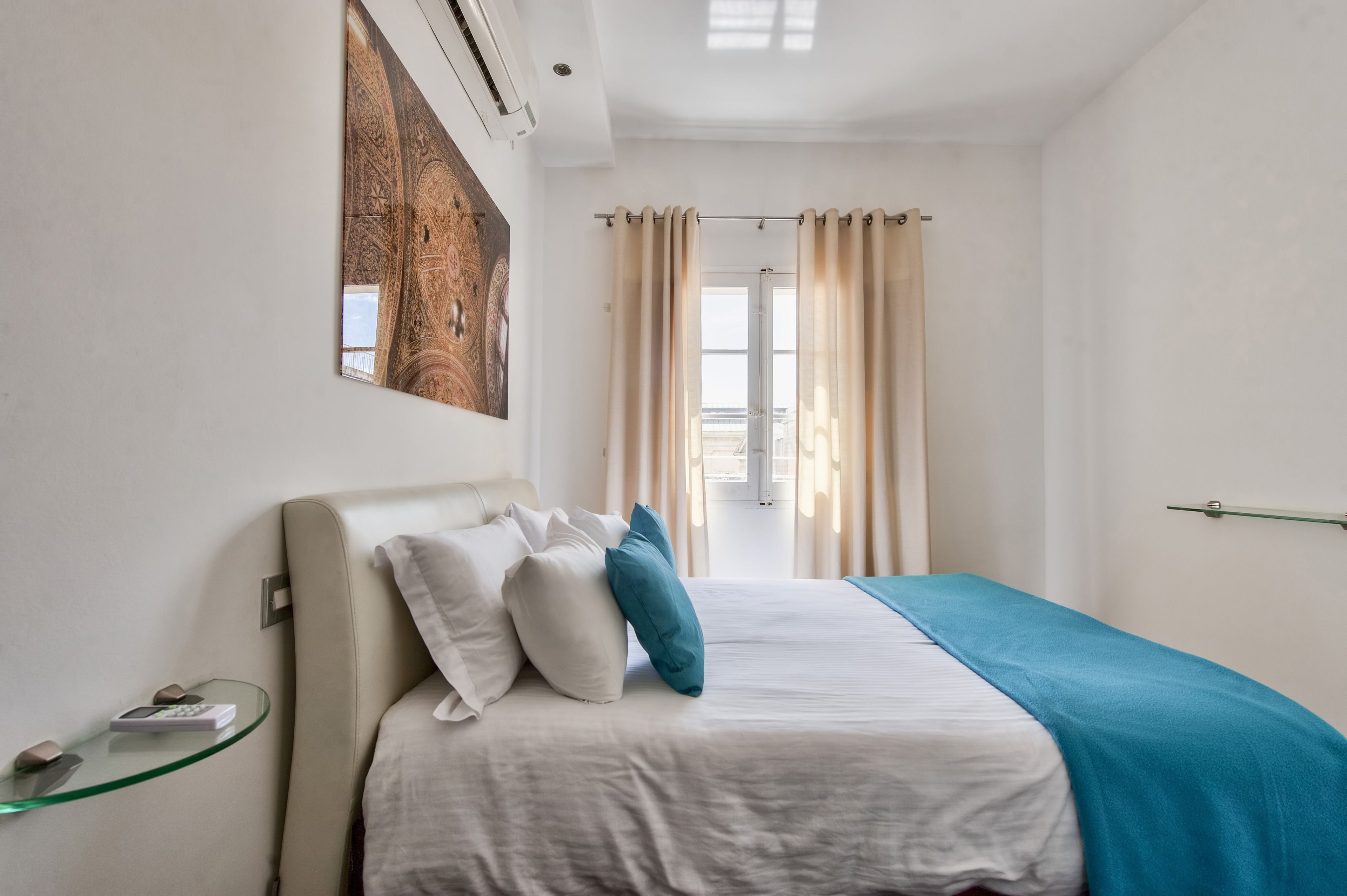 Camera d'albergo con un letto, un tavolino in vetro con un telecomando, un condizionatore d'aria a parete e un'ampia finestra con tende.