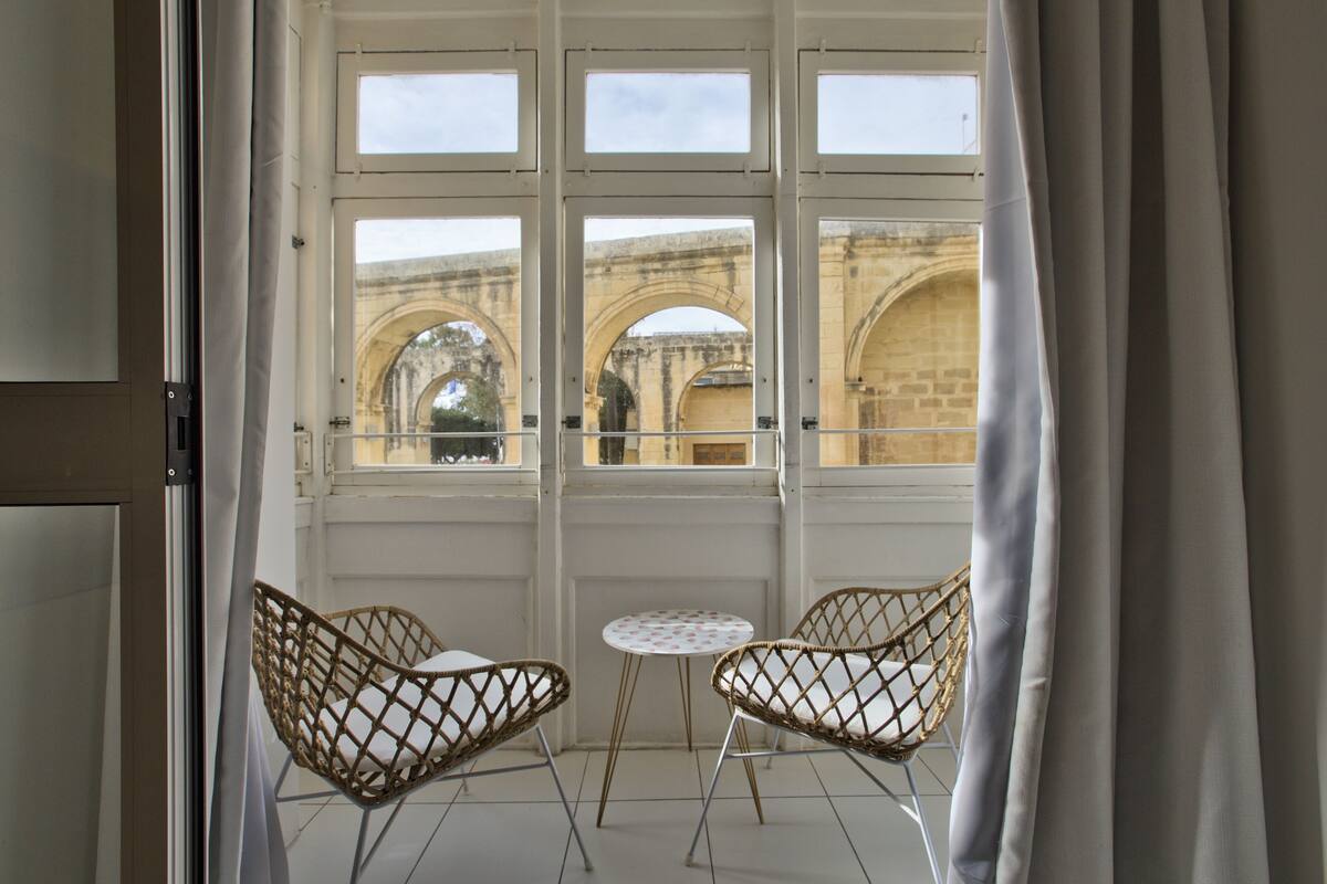 A room with two wicker chairs and a small round table, a large window with multiple panes, and a view of an old stone bridge.