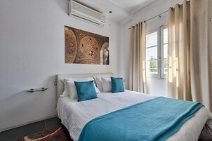 Quarto de hotel com uma cama grande, ar condicionado fixado na parede, janela com cortinas e uma obra de arte decorativa no teto.