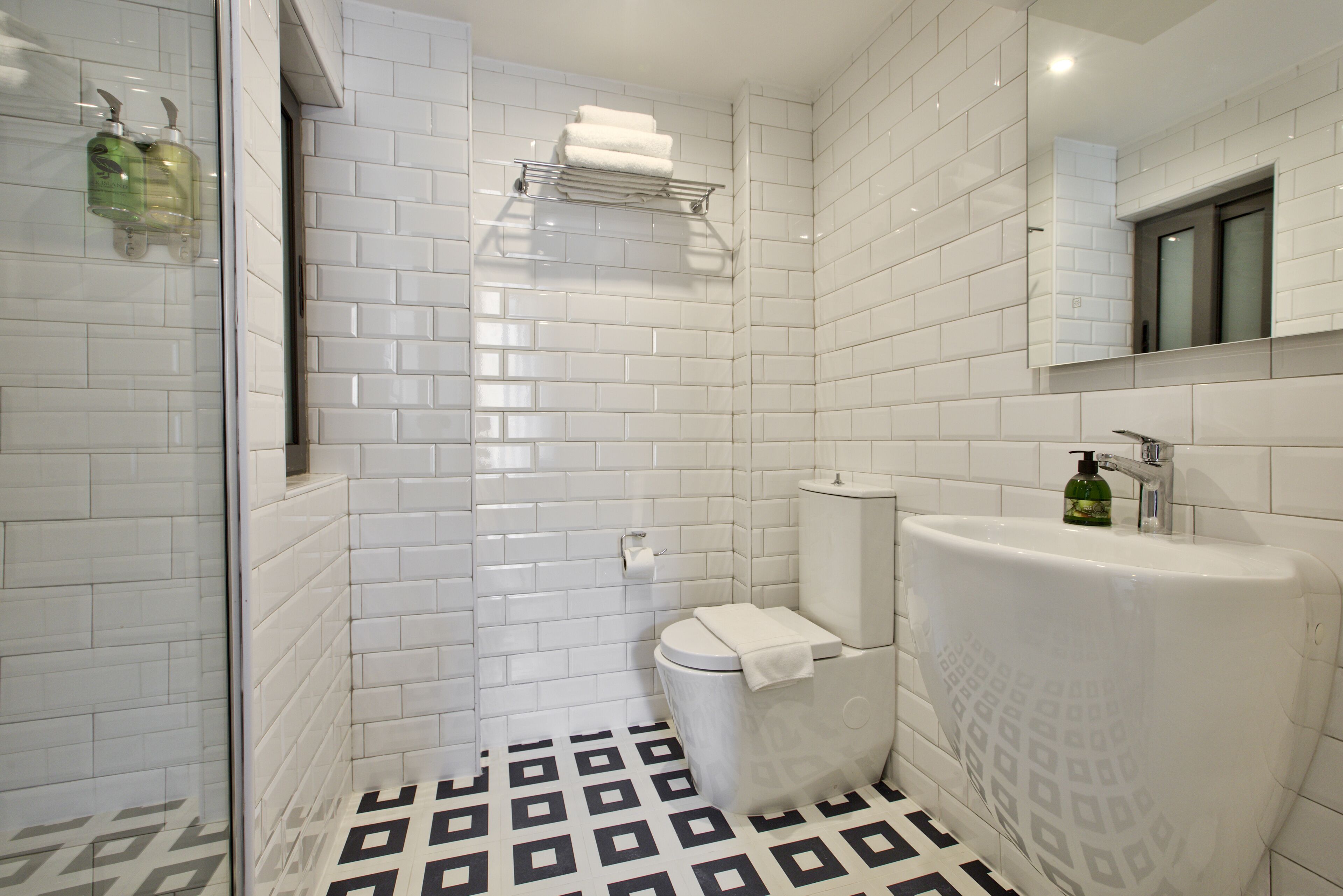 Ein modernes Badezimmer mit weißen, im Fischgrätmuster angeordneten Fliesen, einem schwarz-weißen geometrischen Boden, einer Duschkabine aus Glas, einer weißen Toilette und einer großen, weißen Badewanne.