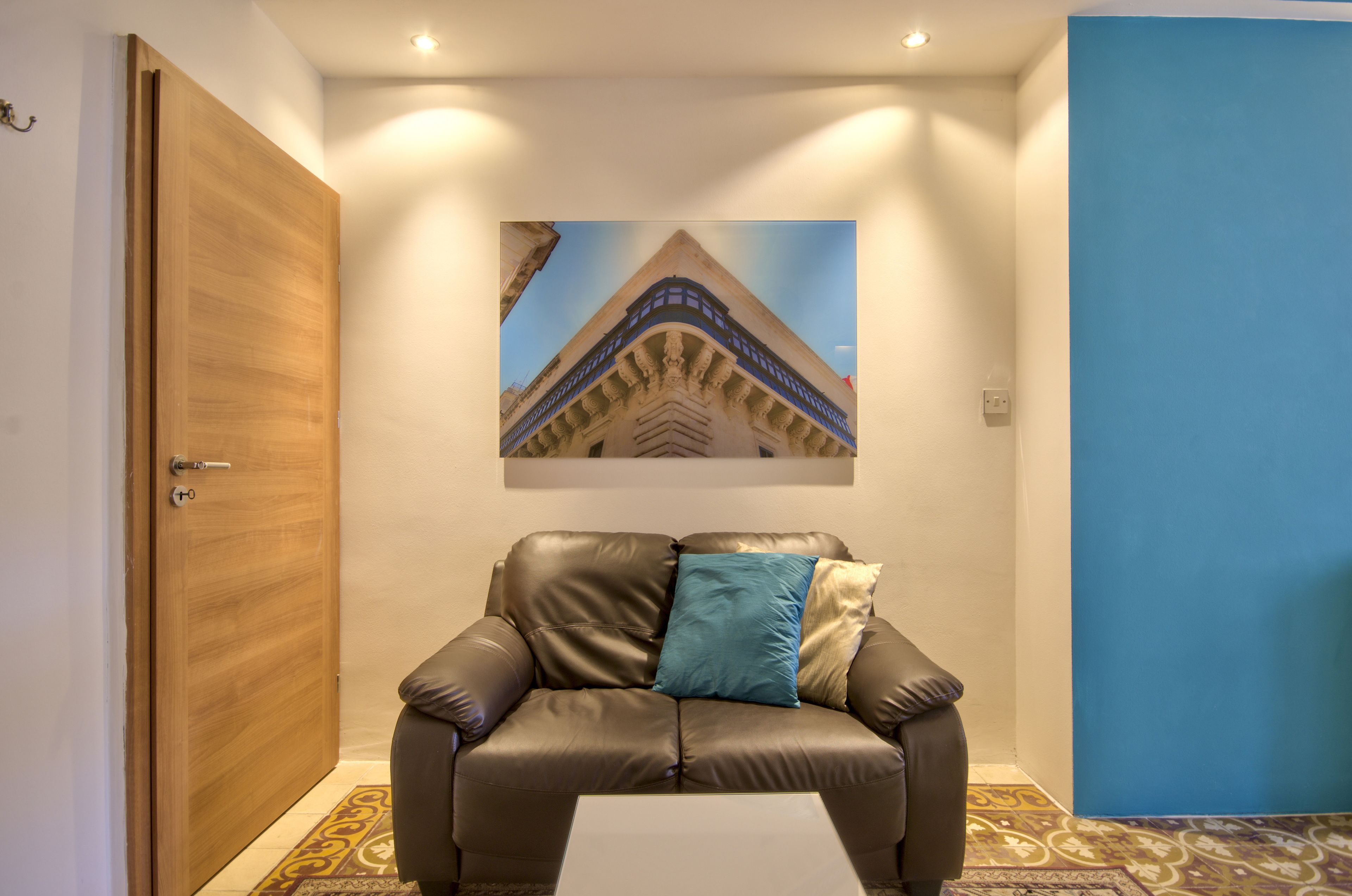 A room with a brown leather sofa, a blue and beige pillow, a framed picture on the wall, and a patterned floor.