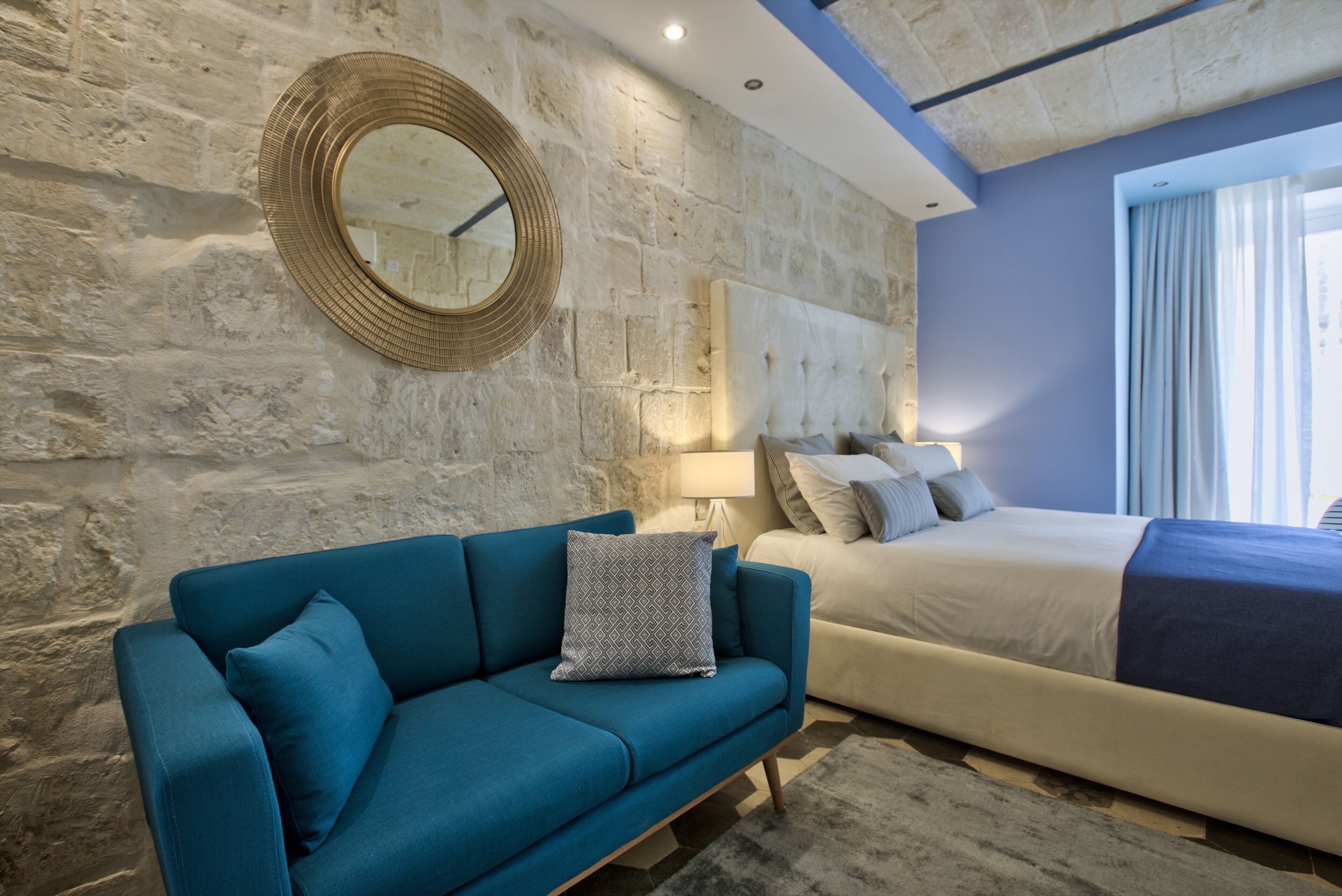 Ein modernes Schlafzimmer mit einer Wand aus Stein, einem runden Spiegel, einem blauen Sofa, einem Bett mit blauer und weißer Bettdecke und einem Nachttisch mit Lampe.