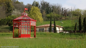 A red glasshouse with a flag on top, a white building with a tiled roof, and a wooden fence in a grassy field. - Hotel Le Foresterie Dei Piaceri Campestri (Varese)