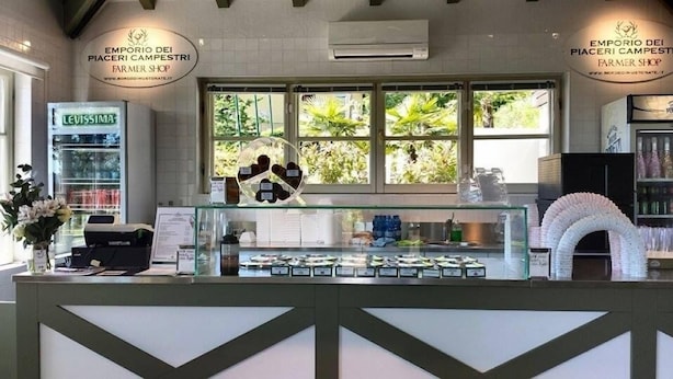A farmer shop with a glass display case filled with products, a cash register, and a sign above the case. - Hotel Le Foresterie Dei Piaceri Campestri (Varese)