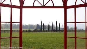 A view through a red-framed window with a geometric design, overlooking a grassy field with a wooden fence and tall trees in the distance. - Hotel Le Foresterie Dei Piaceri Campestri (Varese)