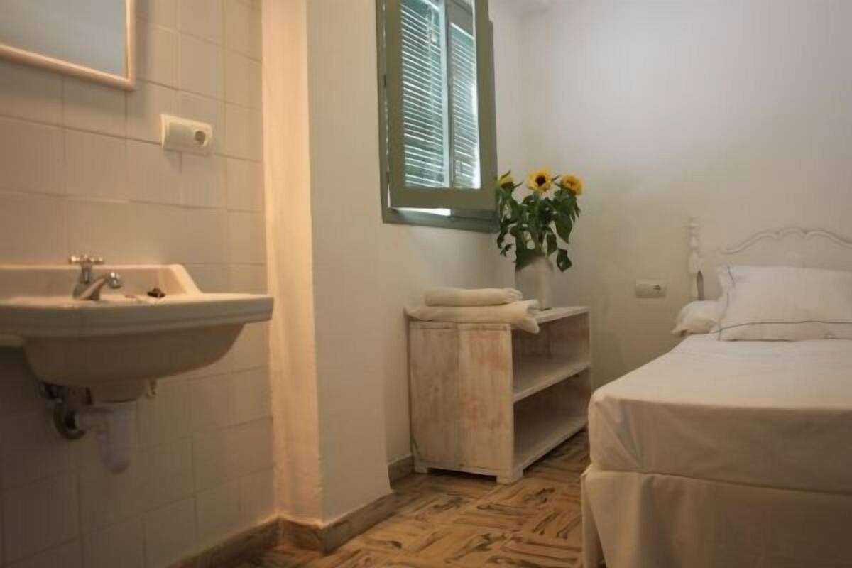 A small, clean room with a single bed, a bedside table, a nightstand, a window with blinds, and a sink.