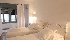Un dormitorio con dos camas, una ventana con contraventanas y una mesita de noche con lámpara.