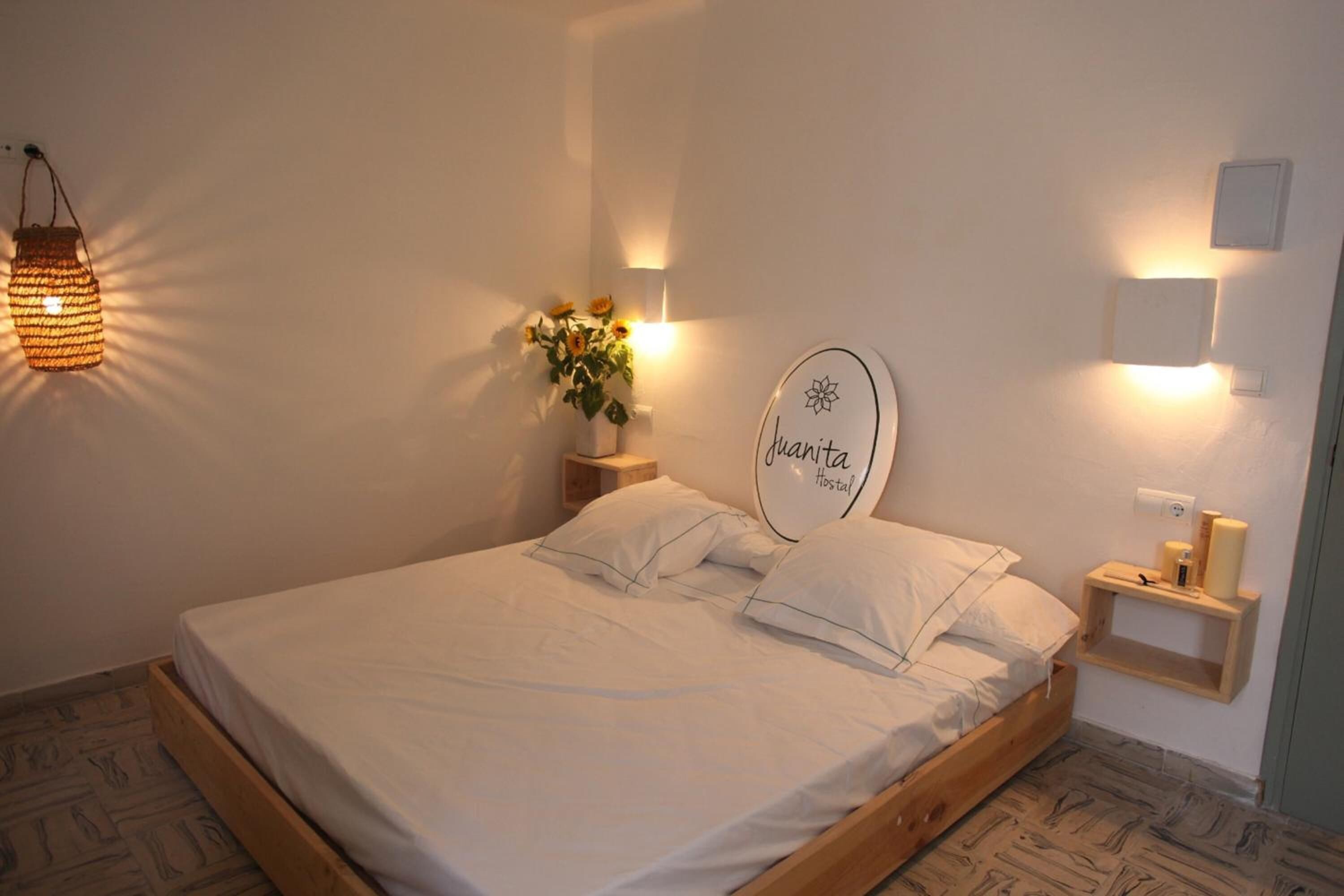 Una cama bien tendida con sábanas blancas, una mesita de noche con lámpara, un letrero en la pared que dice "Granita Hostel" y un estante pequeño con velas.
