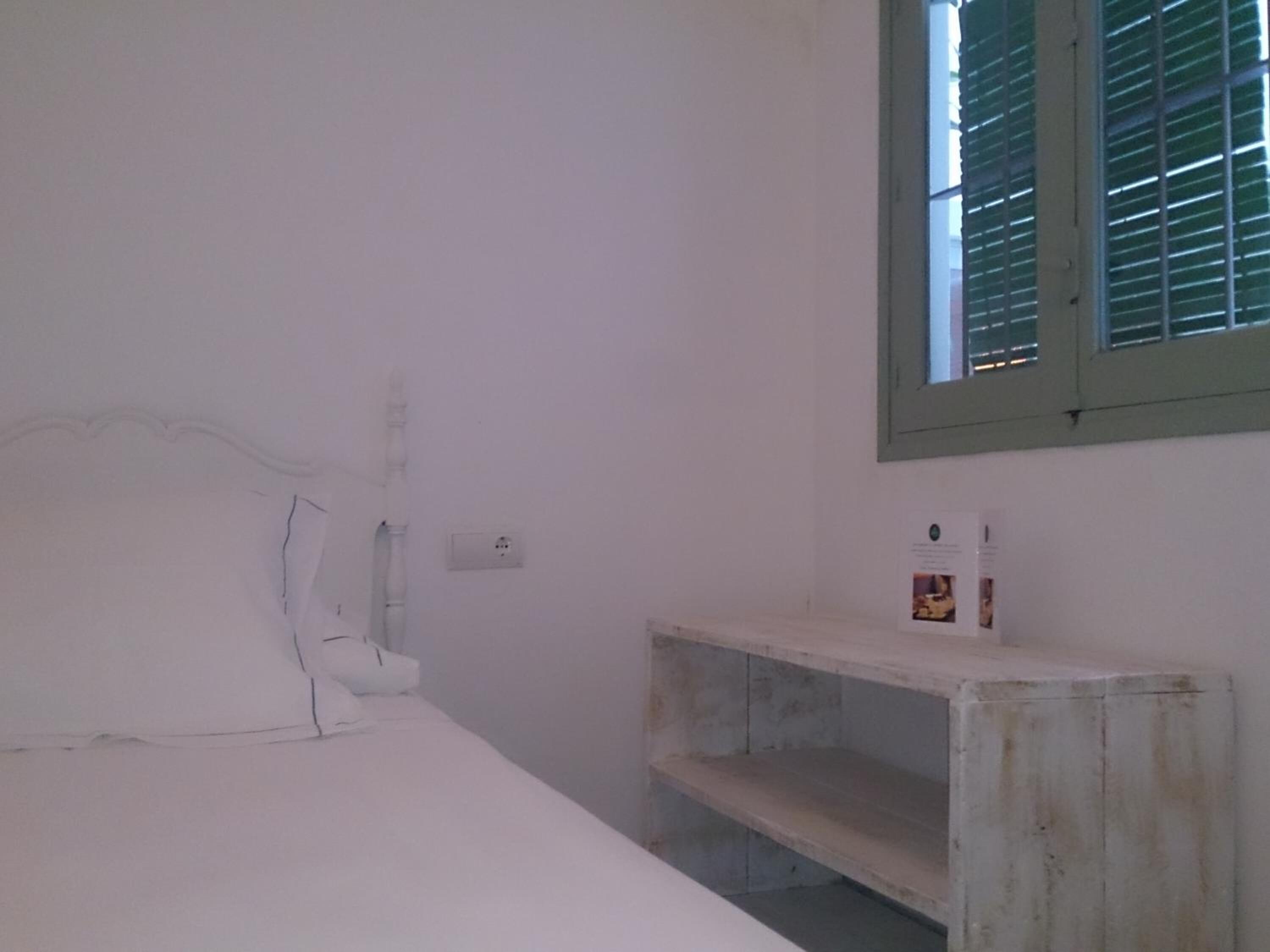 Une pièce comprenant un lit, une petite étagère en bois, une fenêtre avec des volets et une prise de courant au mur.