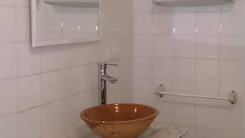 Une salle de bain avec un mur recouvert de carreaux blancs, un lavabo blanc avec un récipient marron, un miroir blanc et un porte-serviettes blanc.