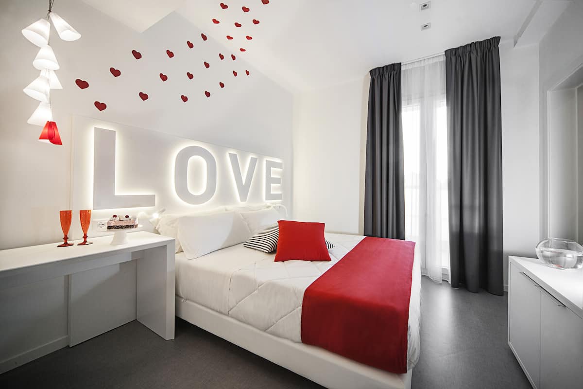 A modern bedroom with a bed, bedside tables, a large illuminated 'LOVE' sign, and a view of the outside through sheer curtains.