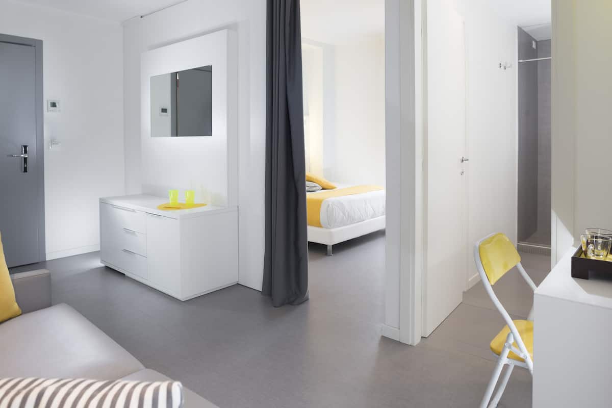 A modern hotel room with a bed, a bedside table with a mirror, a chair, and a small desk area.