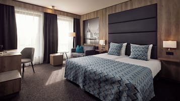 Habitación de hotel con una cama grande, un escritorio, una silla, una mesita y un televisor montado en la pared.