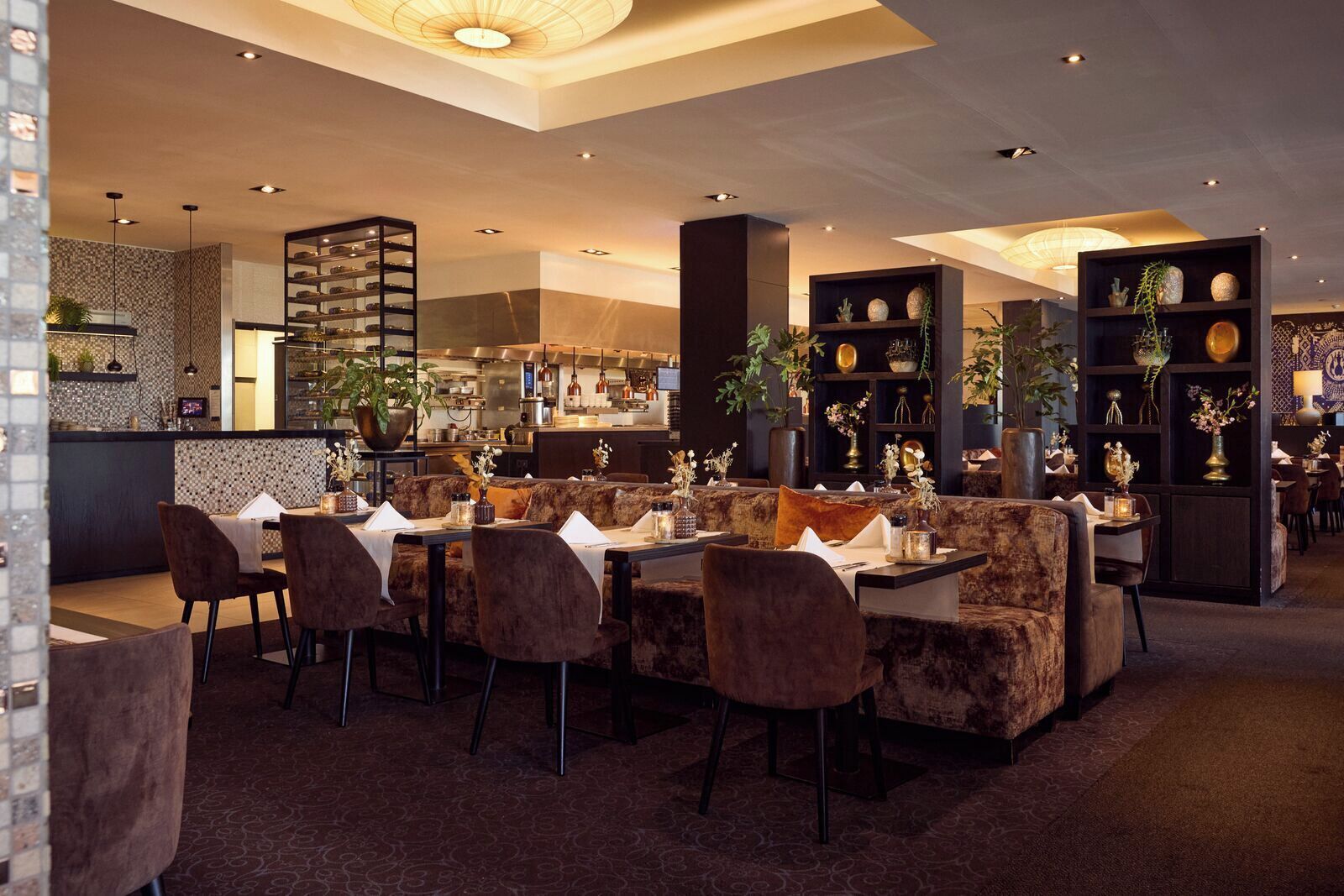 A restaurant with a bar area, seating with velvet chairs, and decorative shelves with plants.