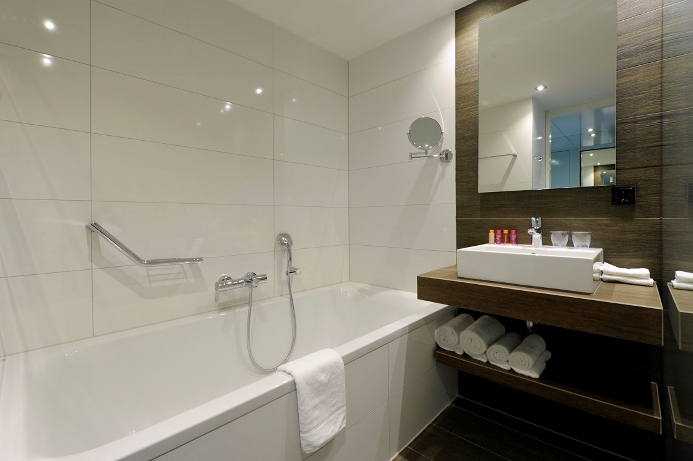 A modern bathroom with a white bathtub, a vanity with a mirror, and a shelf with towels.