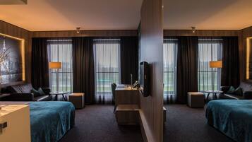 A hotel room with a bed, two sofas, a desk, a TV, and large windows with curtains.