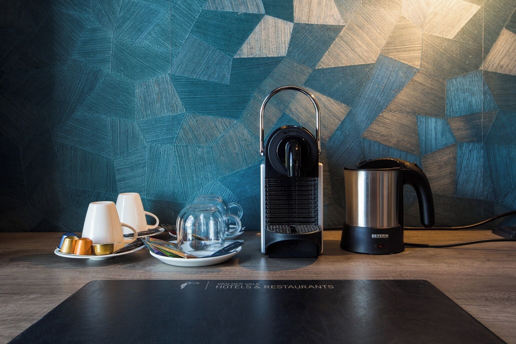A coffee maker, a kettle, and a tray with cups and a glass on a wooden table with a geometric patterned wall in the background.