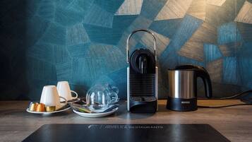 A coffee maker, a kettle, and a tray with cups and a glass on a wooden table with a geometric patterned wall in the background.