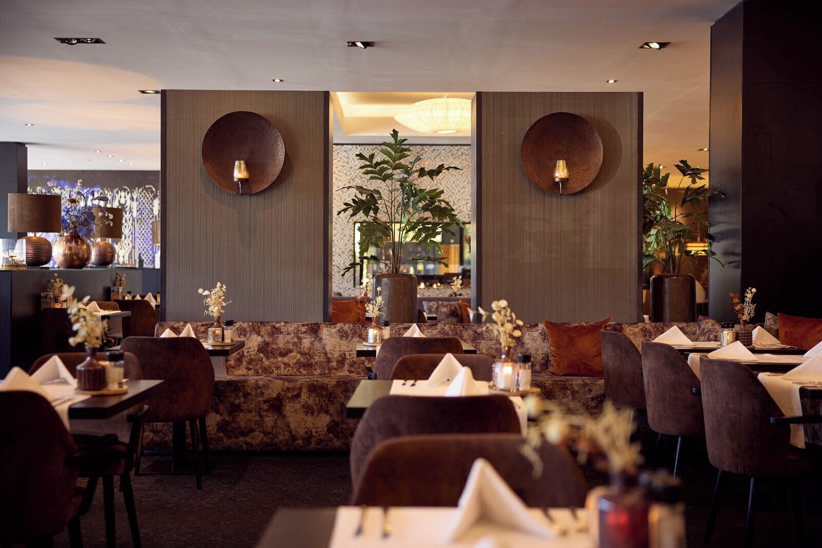 A restaurant with a modern interior, featuring a mix of seating options including tables and a sofa area. There are decorative wall sconces and a large plant in the center.