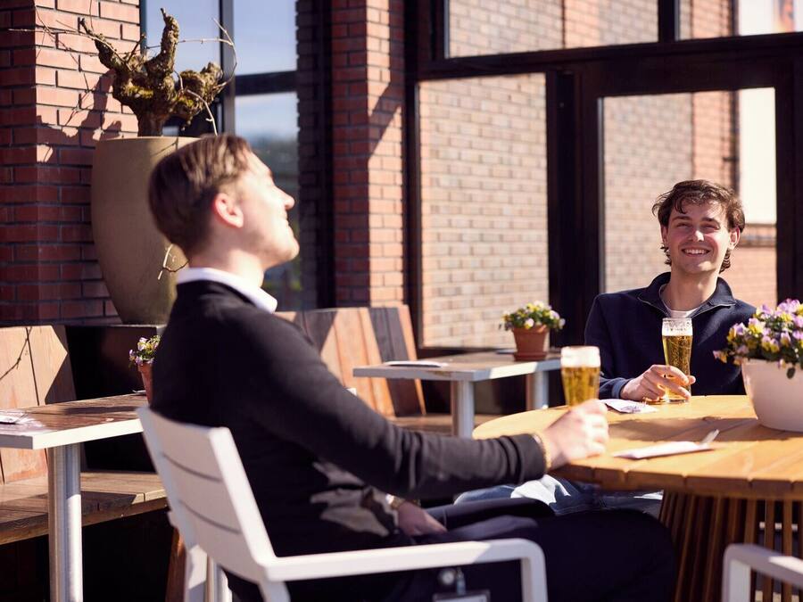Two people seated at a wooden table in an outdoor cafe with brick walls and large windows.