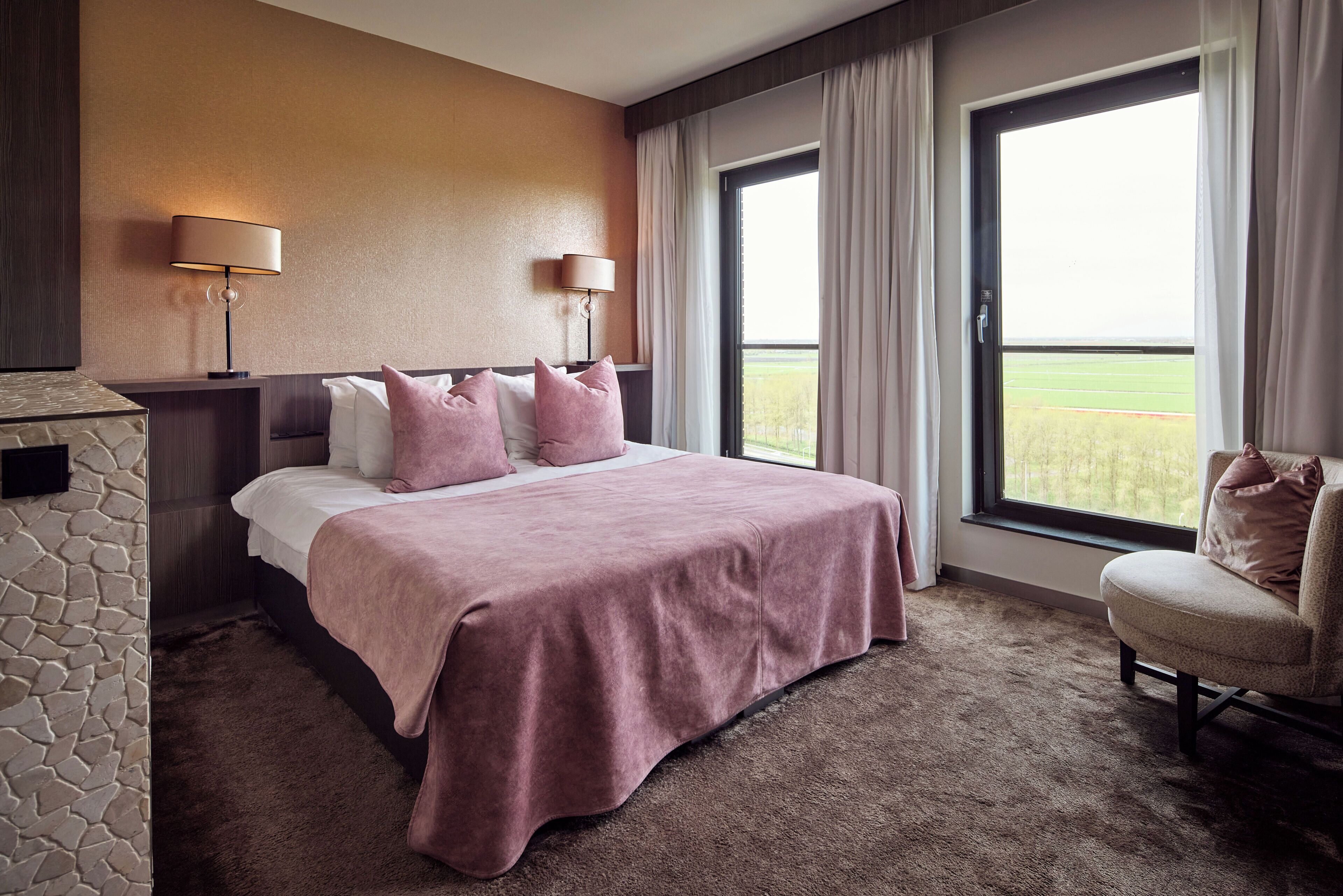 A hotel room with a bed, two bedside lamps, a chair, a stone wall, and a large window with curtains.