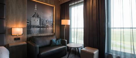 A modern hotel room with a dark leather sofa, a small round table, a single bed, and a large window with curtains.