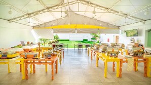 A covered dining area with multiple tables and chairs, a tented roof, and a view of a green outdoor space.