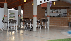 Un patio al aire libre con sillas y mesas de metal, una pared de ladrillo con una zona de bar y una piscina al fondo.