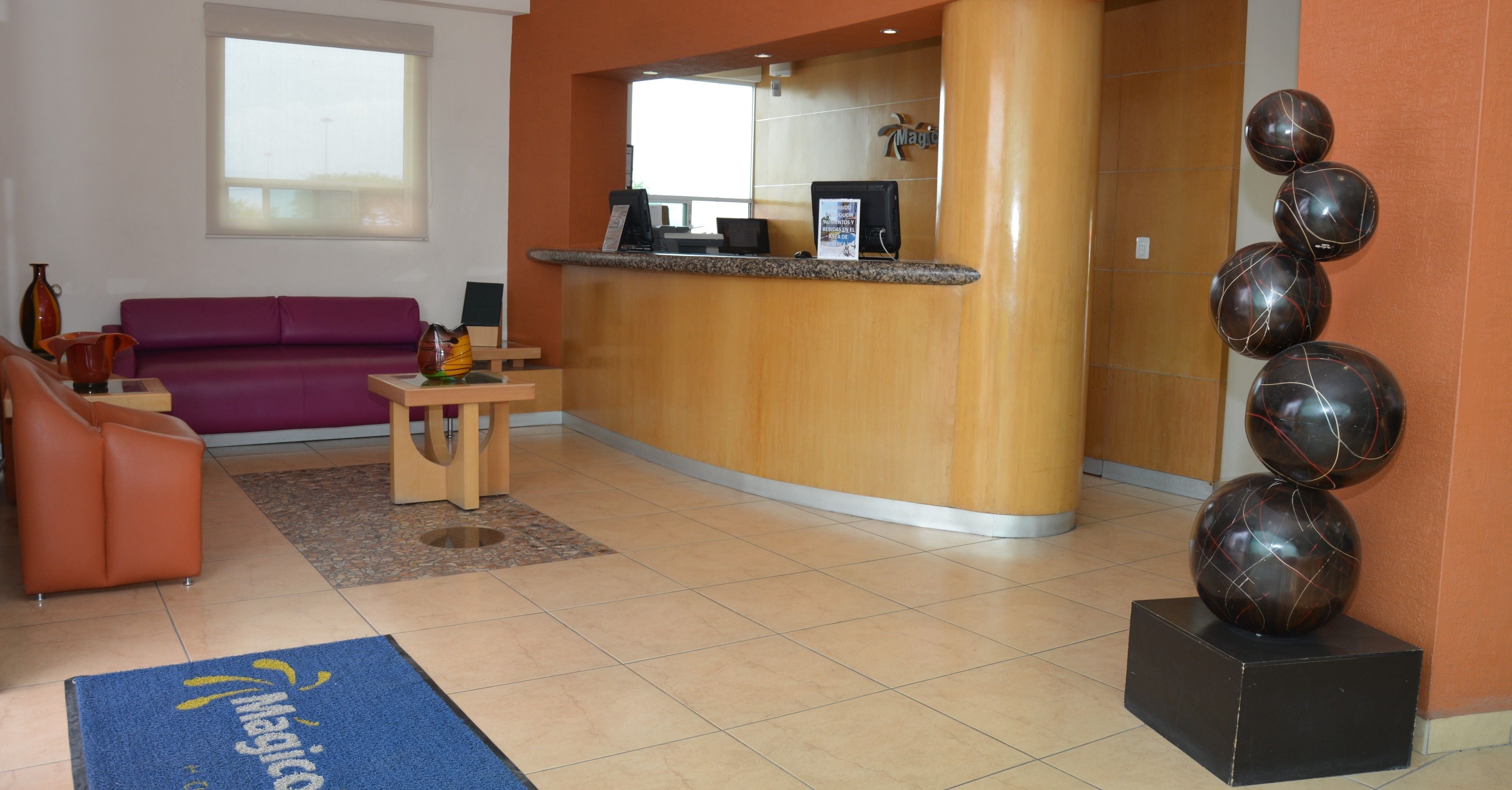 Een hotellobby met een receptiebalie, een paarse bank, een kleine tafel met een vaas en een blauwe deurmat.