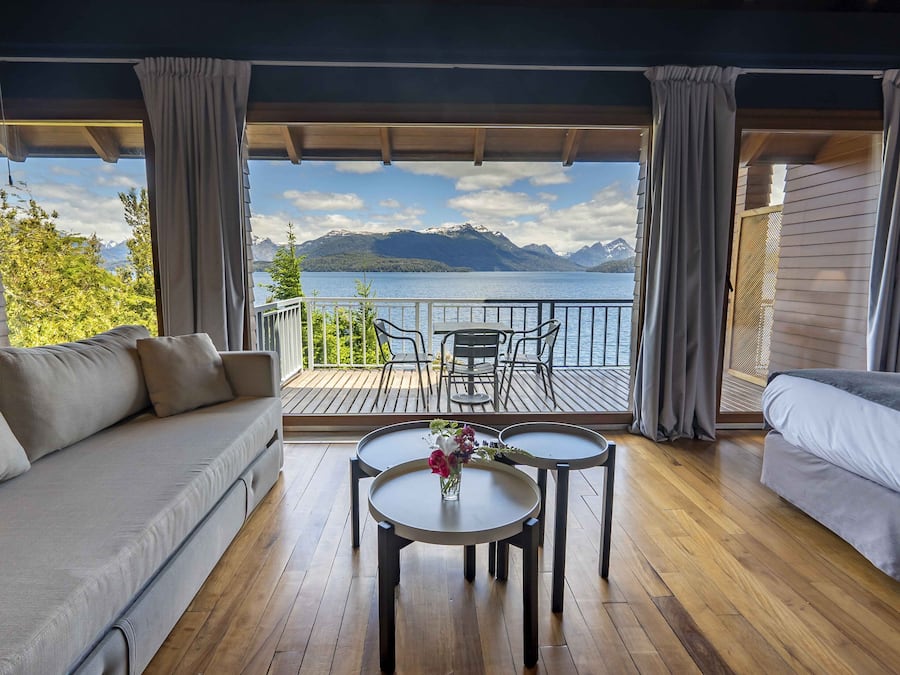 A room with a sofa, a small table with a vase of flowers, and a view of a lake and mountains.