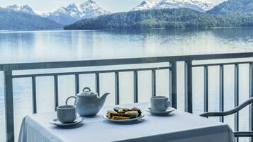 Una mesa con un calentador de agua, tazas y un plato de pasteles en un balcón con vistas a un lago y montañas al fondo.