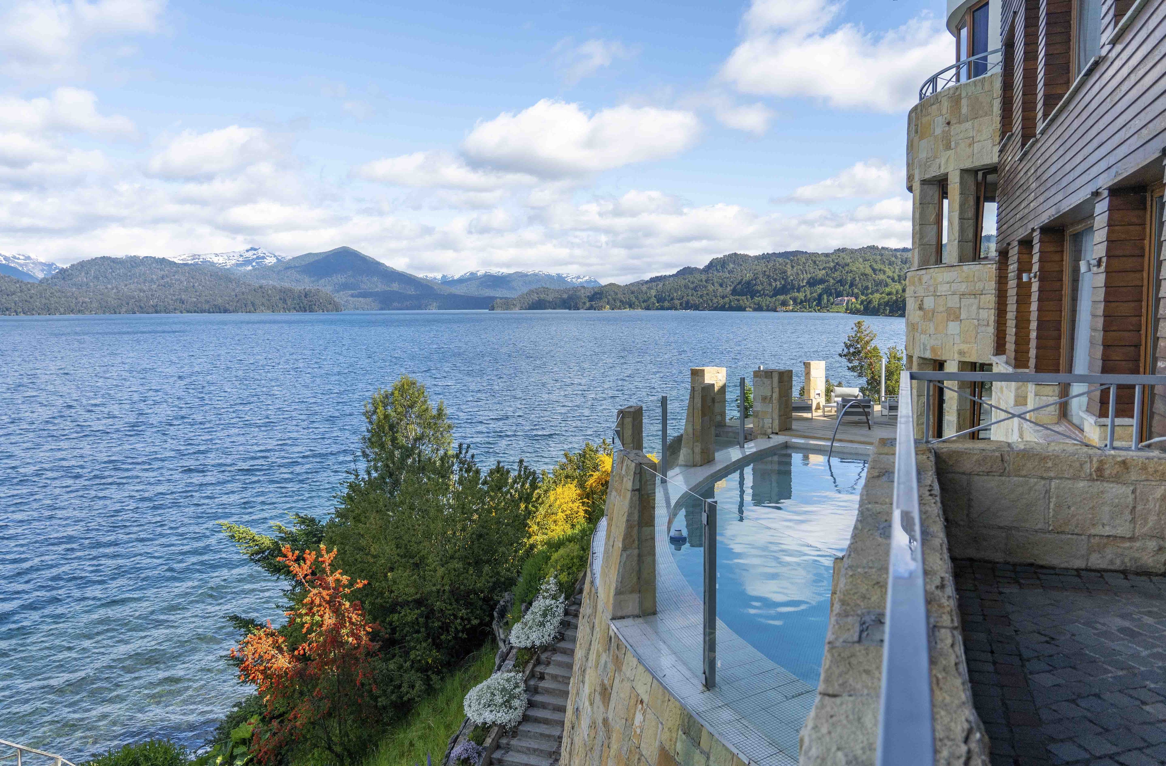 Un edificio moderno con piscina infinita de cristal con vistas a un lago, arquitectura de piedra y montañas al fondo.