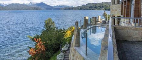 Un edificio moderno con piscina infinita de cristal con vistas a un lago, arquitectura de piedra y montañas al fondo.