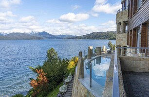 Un edificio moderno con piscina infinita de cristal con vistas a un lago, arquitectura de piedra y montañas al fondo.