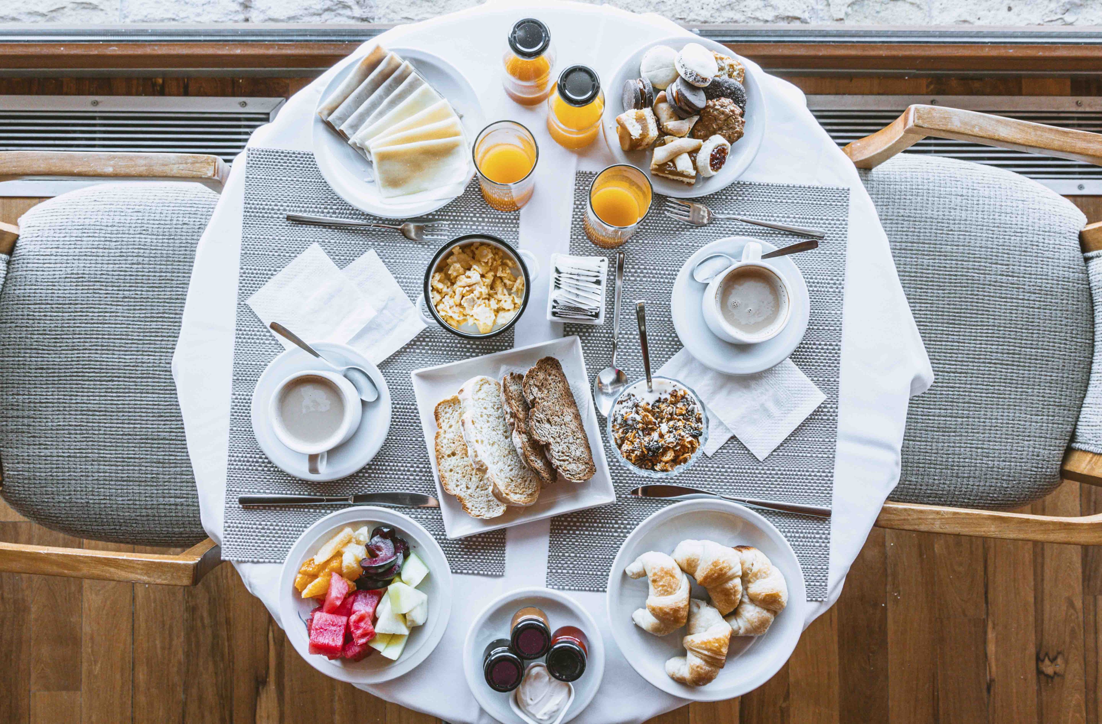 A round table set with breakfast items including bread, fruits, and beverages, surrounded by grey upholstered chairs.