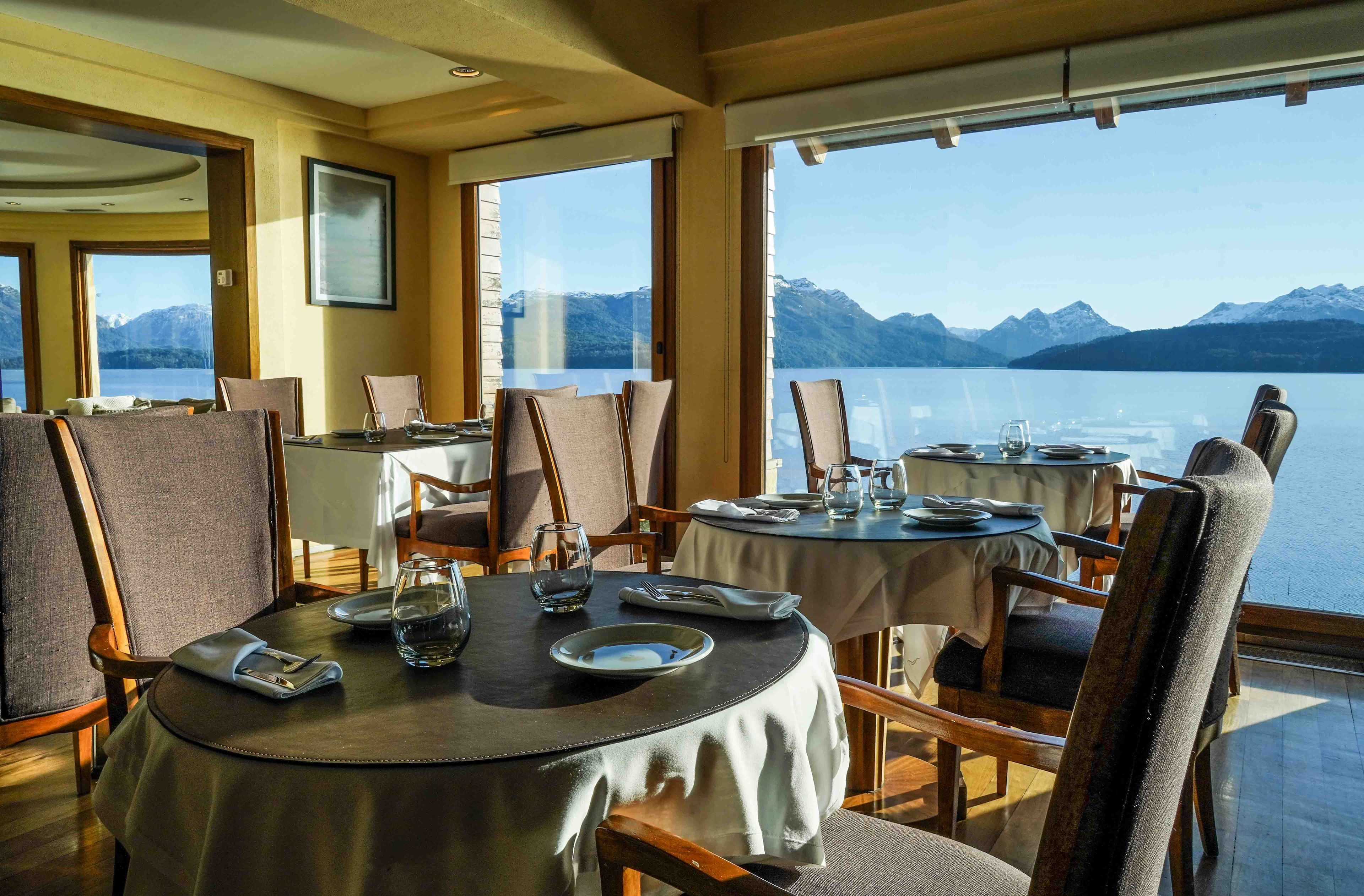 Un espace repas avec des tables rondes, des chaises et une vue sur un lac et des montagnes.