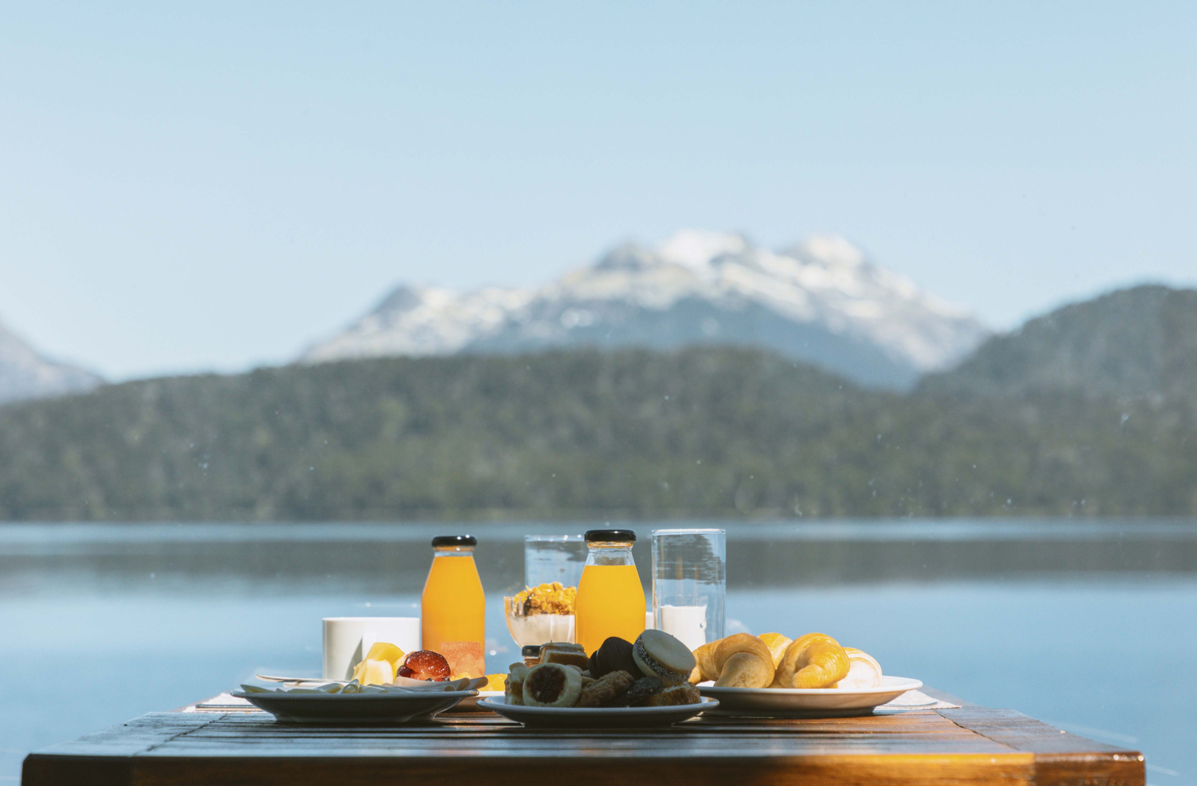 A breakfast table with various food items and drinks, set against a backdrop of a lake and mountains.