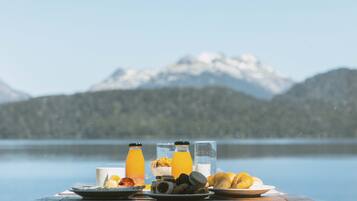 A breakfast table with various food items and drinks, set against a backdrop of a lake and mountains.