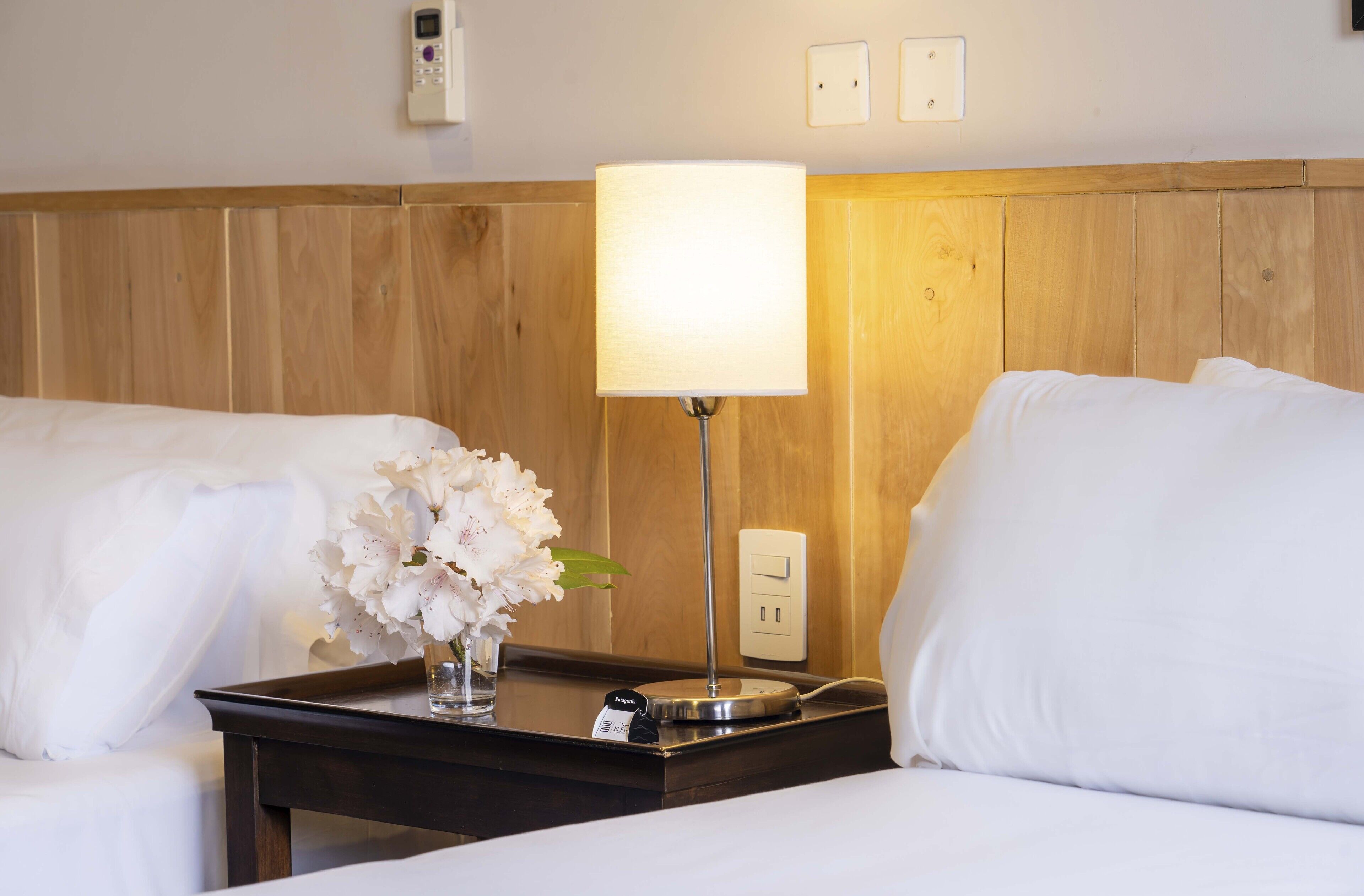 A hotel room with two beds, a bedside table with a lamp, a vase with flowers, and a power outlet.