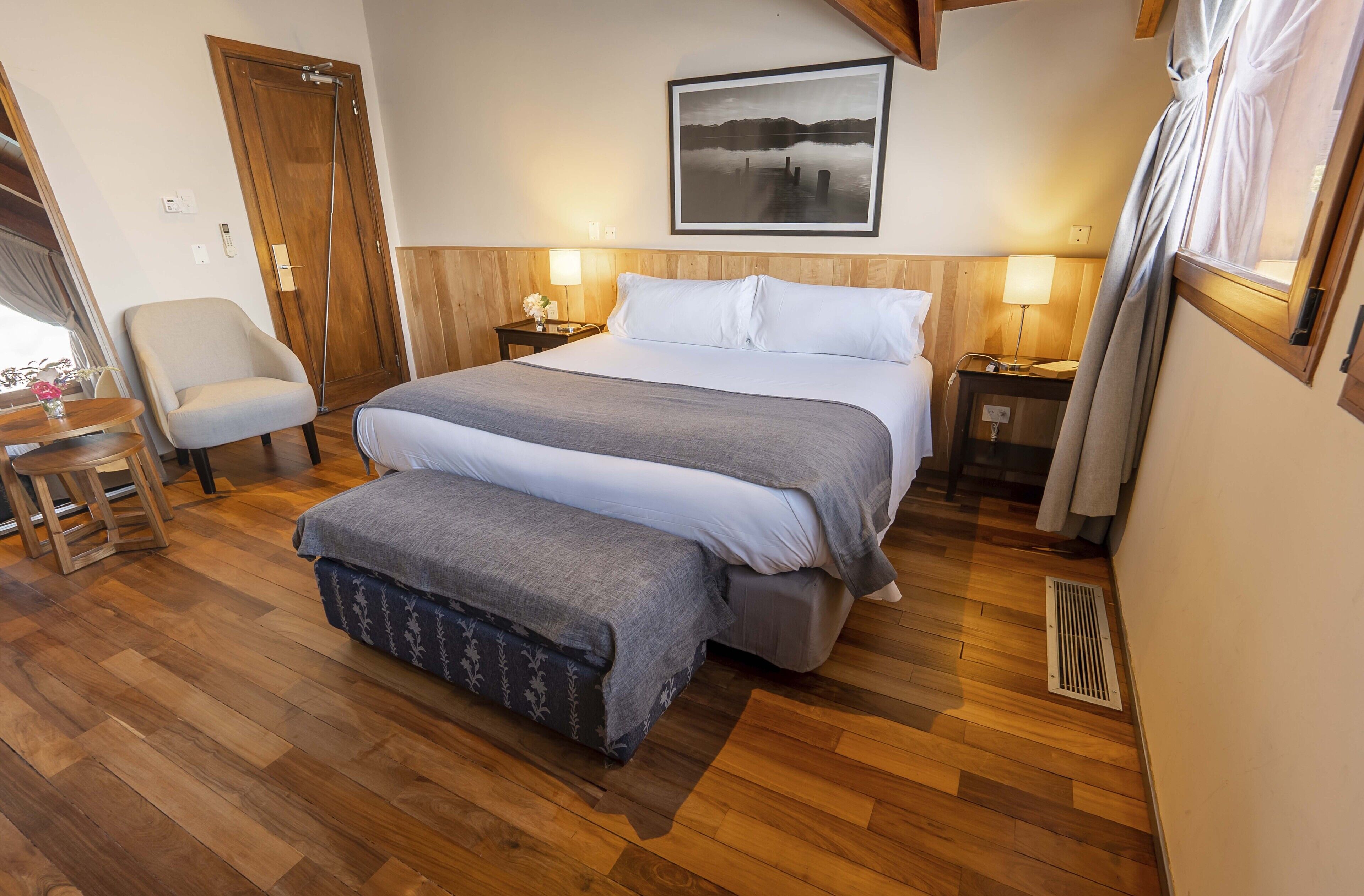 Un dormitorio con una cama grande, una silla, una mesita de noche con lámpara y suelo de madera.