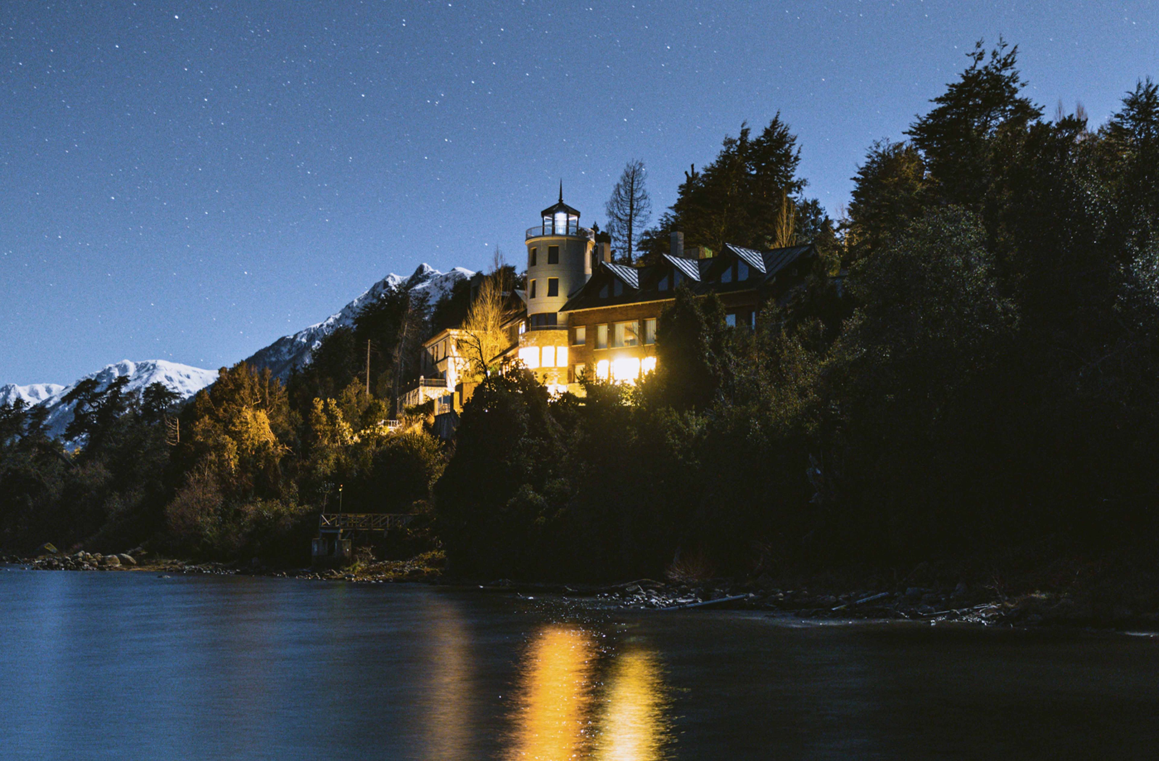 Un edificio grande e iluminado con una torre, rodeado de árboles y montañas nevadas, que se refleja en el agua.
