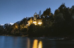 A large, illuminated building with a tower, surrounded by trees and snow-capped mountains, reflecting on a body of water. - El Faro Boutique Hotel & Spa by DON (Villa La Angostura)